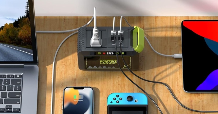 a black and green portable power station on a desk with a laptop, smart phone, Nintendo DS, and table plugged into it charging