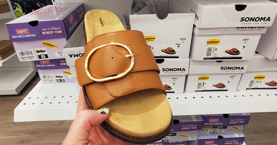 hand holding up a brown leather sandal in front of shoe boxes with clearance stickers