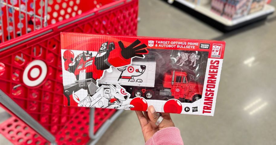 hand holding a Transformers Target Optimus Prime and Autobot Bullseye Action Figure Set in front of Target cart