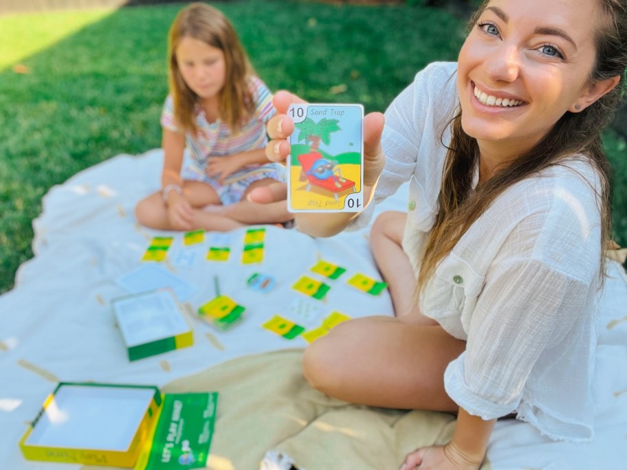 woman smiling and holding a Play Nine card to the camera, with her daughter sitting behind her as they are playing the game on a blanket outdoors