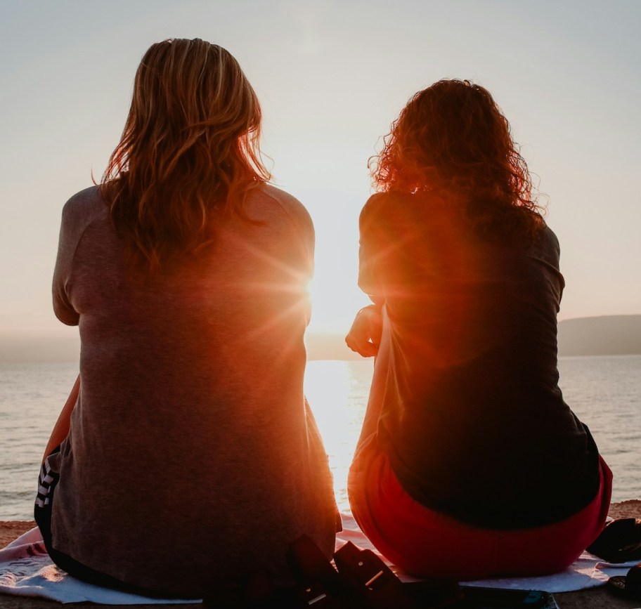 two women sitting on towel looking out at ocean sunset