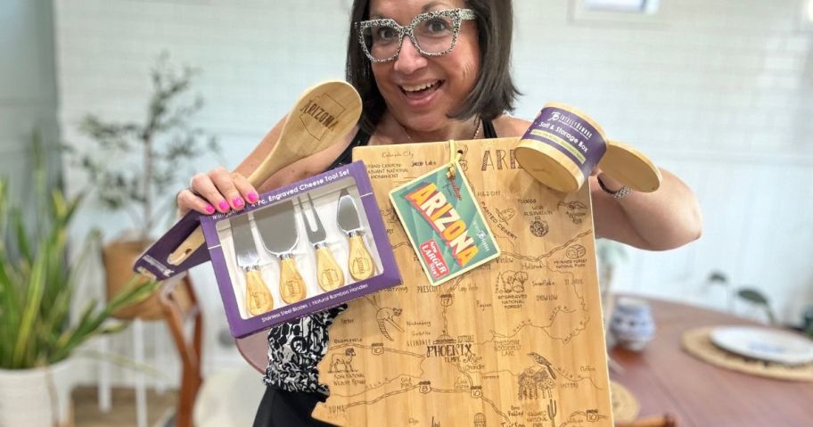 Woman holding up a Bamboo Cutting Board shaped like Arizona along with a set of cheese knives, a spoon and a salt cellar