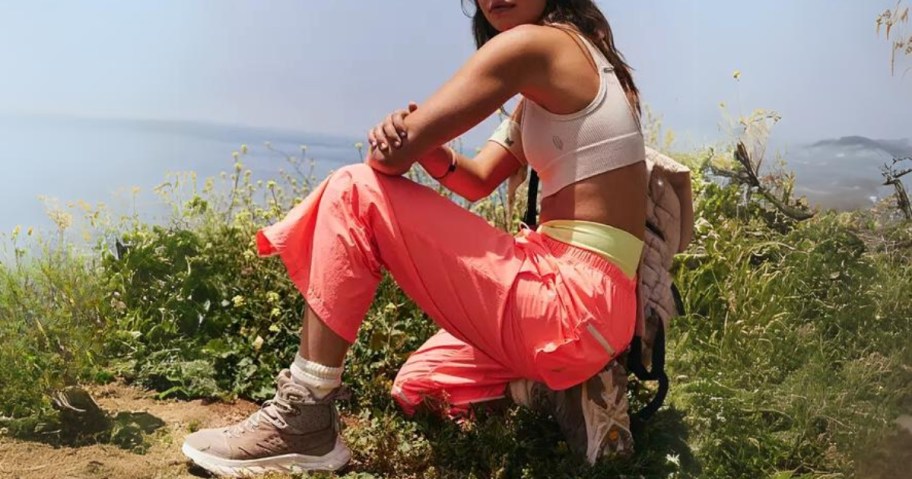 woman squatting outside wearing orange and yellow pants, tan hiking boots, and a white crop top