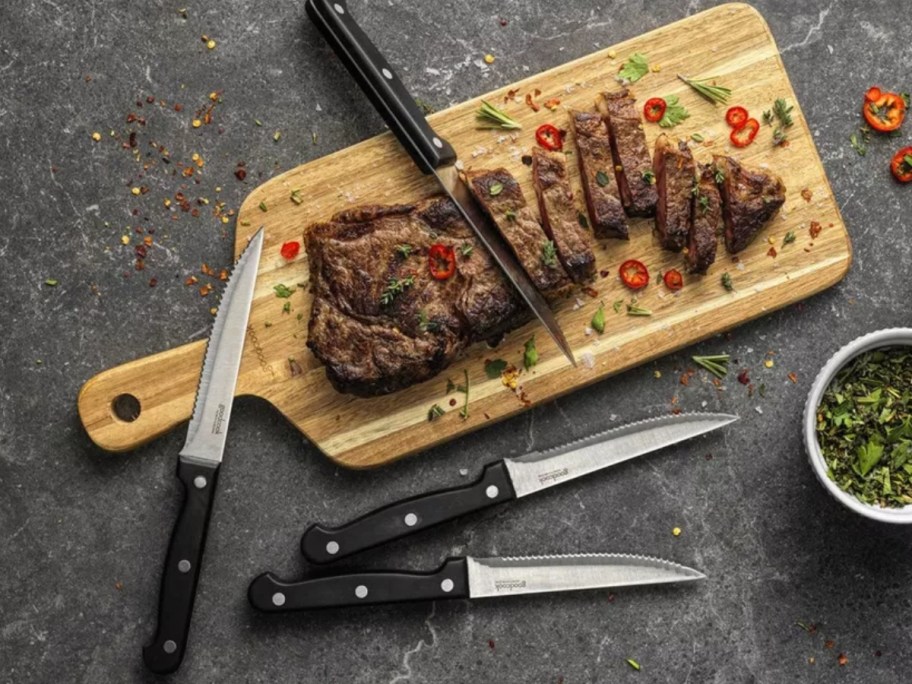 four steak knives with black handles around a wood cutting board that has a sliced steak on it