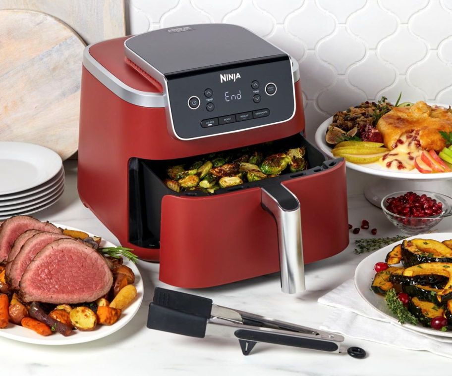 a red ninja air fryer sitting on a white marble countertop shown with dishes of prepared food