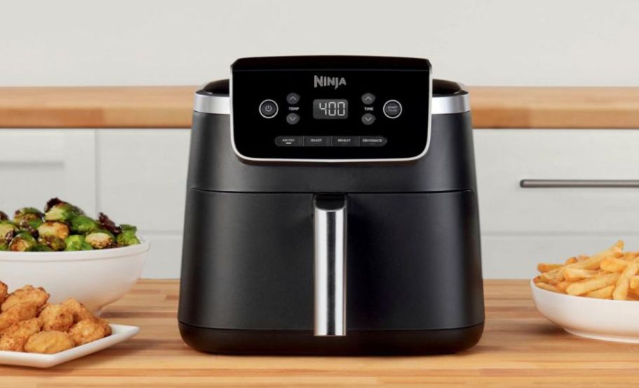 a black ninja airfryer sitting on a butcher block countertop shown with dishes of prepared food