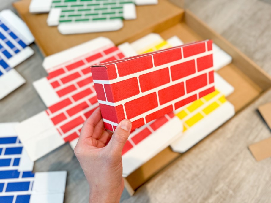 hand holding up an assembled cardboard building block in red, with more unassembled ones in other colors behind it