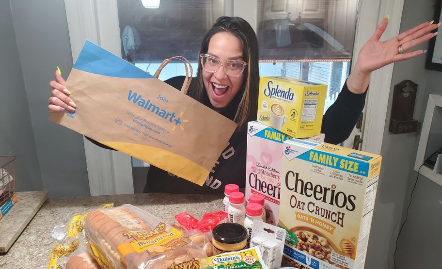 a woman in a kitchen with groceries on the counter holding a walmart plus paper shopping bag and waving