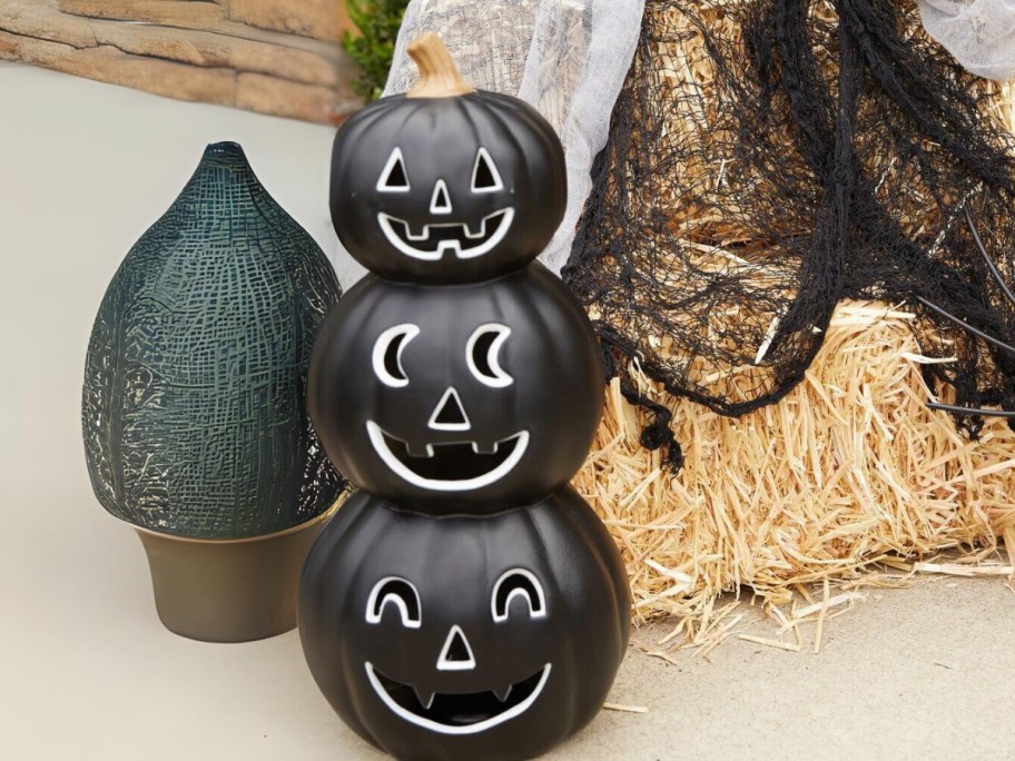 a Halloween decoration stack of 3 black jack-o-lanterns next to a hay bale and other Halloween decor