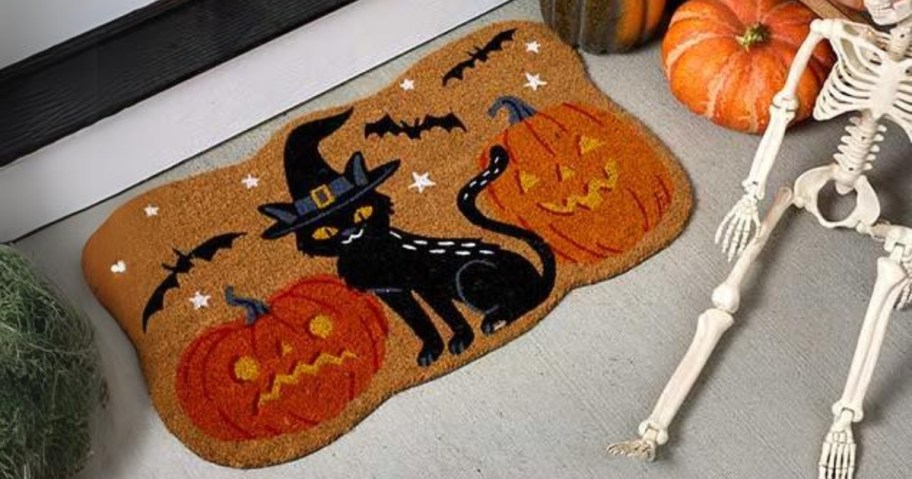 a Halloween coir doormat with a black cat and jackolanterns outside a door next to a skeleton