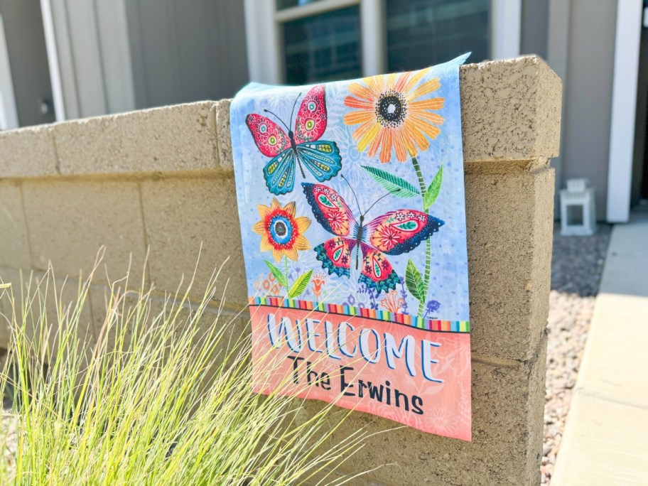 personalized garden flag with butterflies and flowers hanging on a stone fence