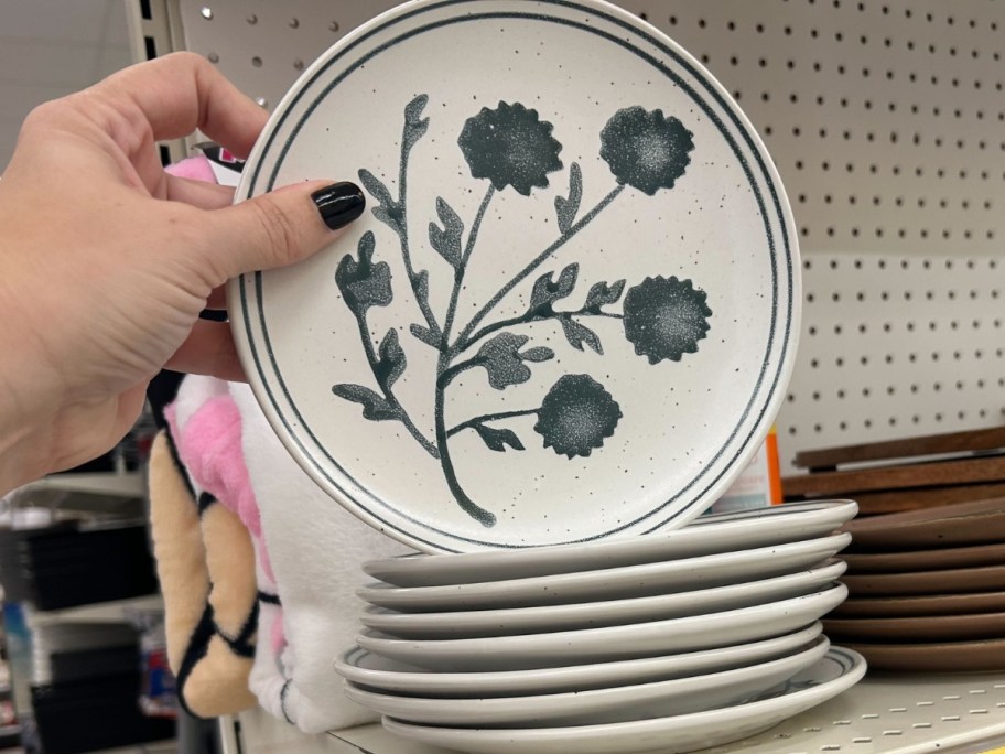 hand holding a white dinner plate with blue flowers, more stacked behind it on a store shelf