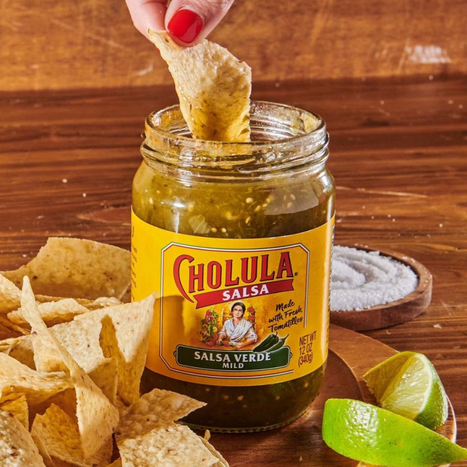 a womans hand dipping a tortilla chip into a jar of salsa verde