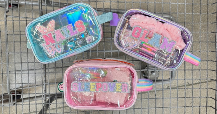 3 clear accessory bags with pink, purple, and blue accents and block letters on them, filled with various accessories in a shopping cart. One says "Glam", one says "Nails" and one says "Sleepover"