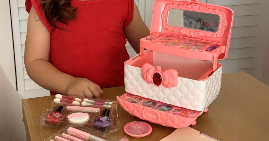 little girl in a red shirt next to a pink and white makeup case with brushes and makeup around it