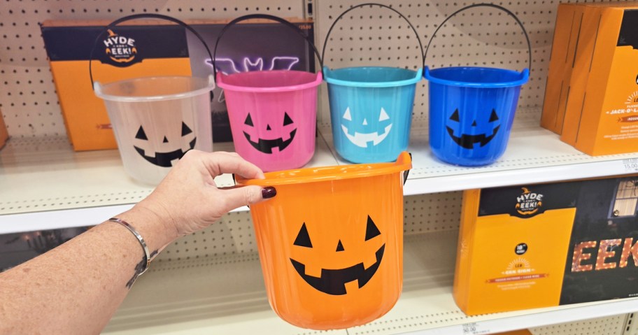 hand holding up an orange pumpkin pail in front of a shelf with other color pails