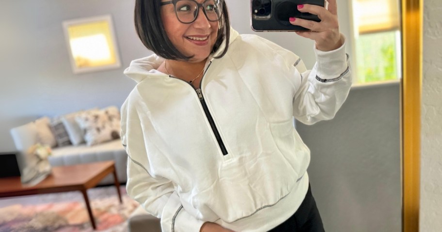 woman wearing a white half zip hoodie cropped sweatshirt taking a selfie in a mirror