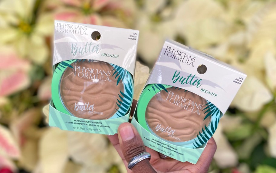 Hand holding two bronzer compacts in packaging near poinsettia flowers 