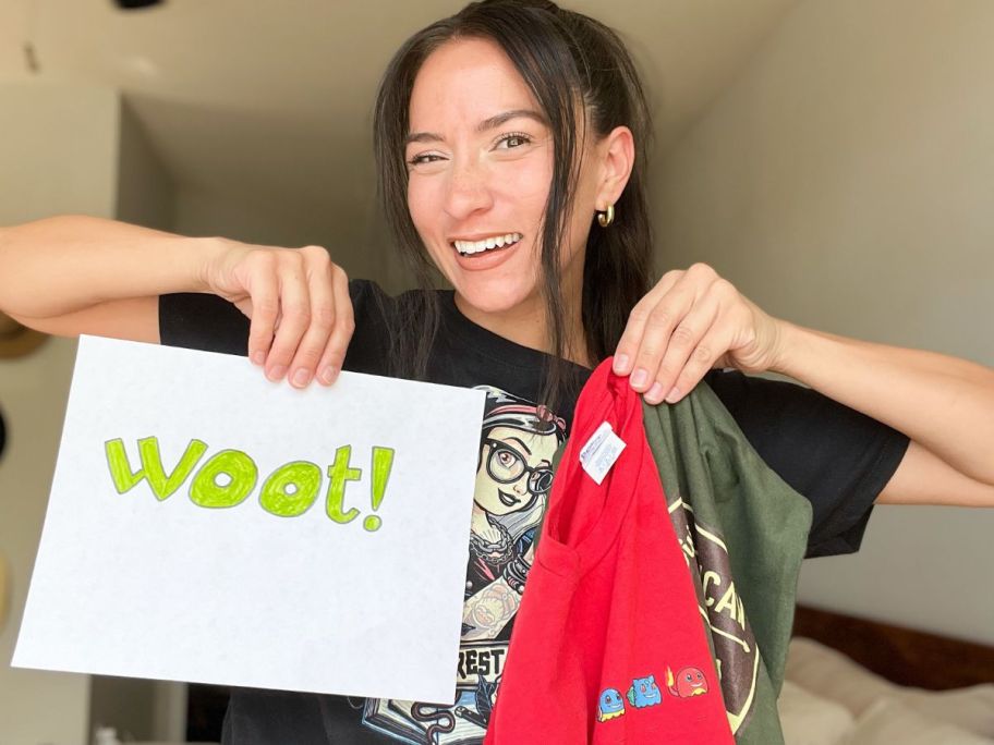 woman holding a paper with WOOT! on it in one hand and two t-shirts in another