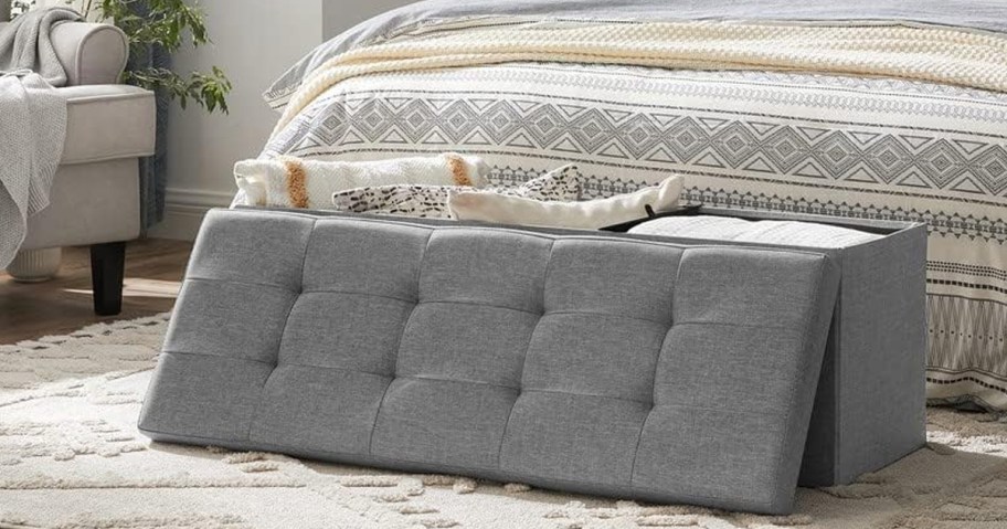 gray ottoman with pillows and blankets with lid laying next to it 