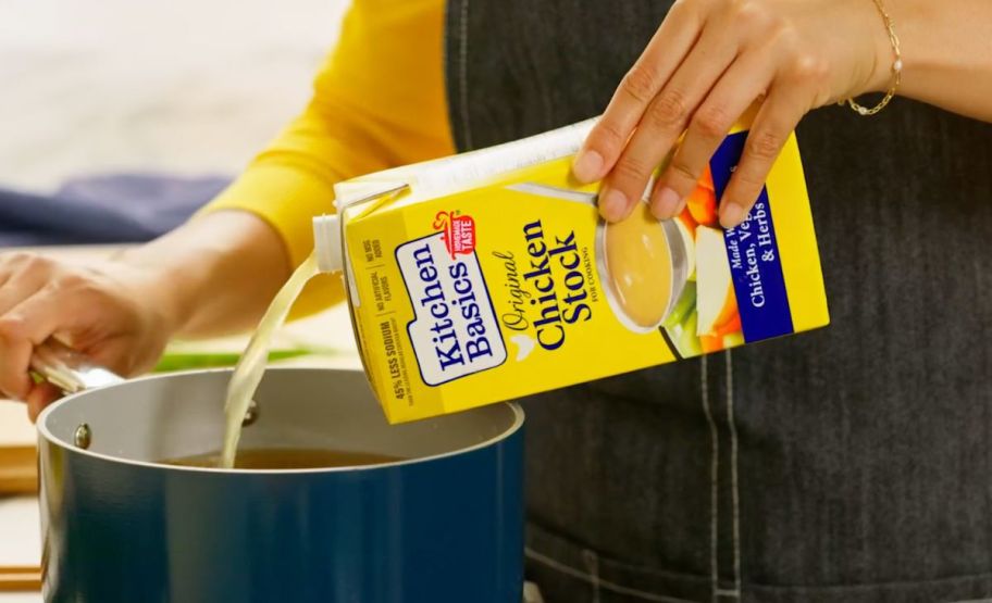 a womans hand shown pouring chicken stock out of a container and into a cooking pot