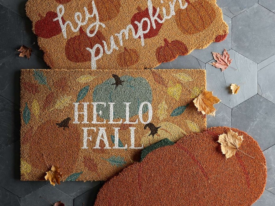 Fall themed coir doormats on a stone floor, one with pumpkins that says "Hey Pumpkin", another with pumpkins that says "Hello Fall" and another orange pumpkin shaped doormat