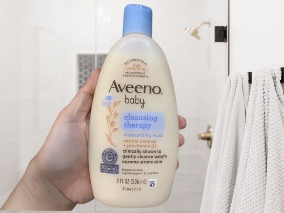 hand holding Aveeno Baby Cleansing Therapy Moisturizing Baby Body Wash 8oz in bathroom