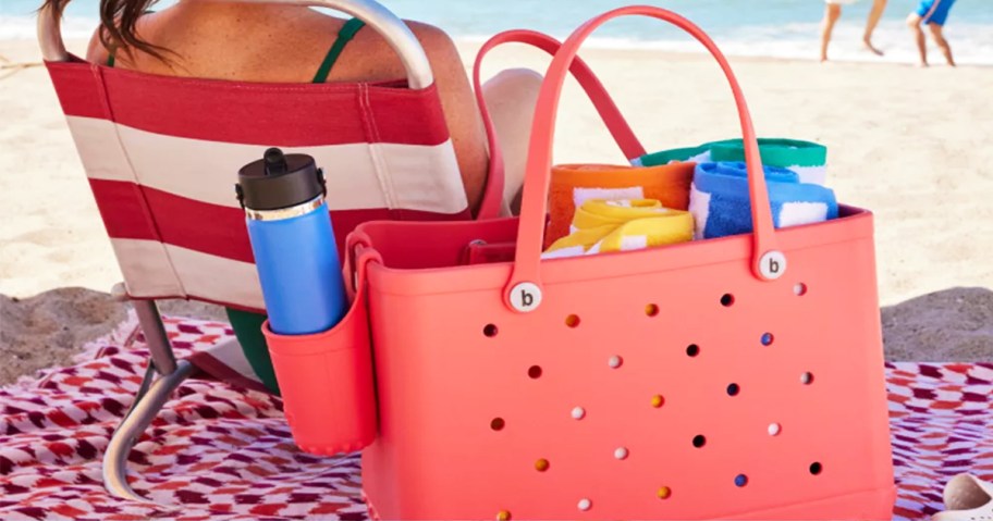 woman sitting on beach with a bight pink bogg bag behind her