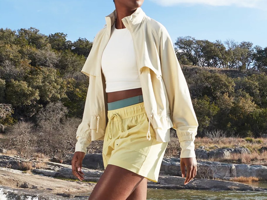 woman walking on beach in white jacket and tank with yellow shorts