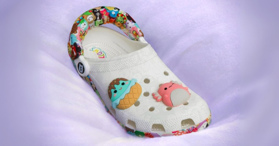  a white Crocs shoe with Squishmallow Jibbitz, multi color sole and cushioned heel strap with various Squishmallows on it, sitting on a purple cushion