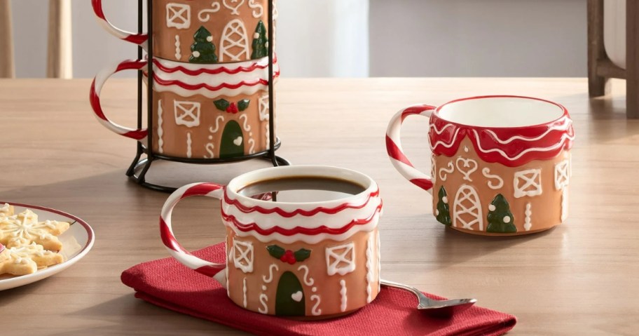 a set of gingerbread house holiday coffee mugs on a table, one with cocoa in it, one empty, 2 stacked in a mug rack behind it