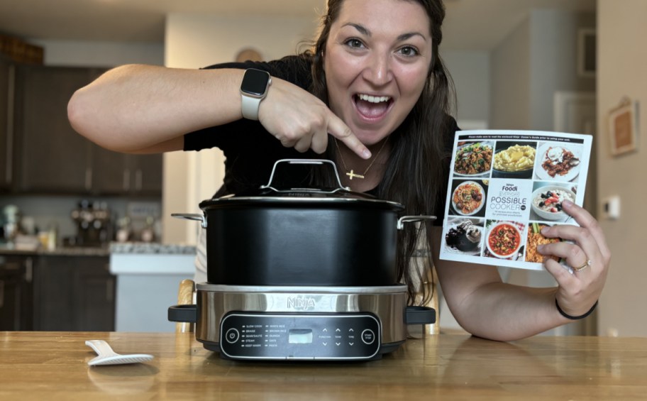 Woman holding a cookbook and pointing to a Ninja Cooker on the counter 