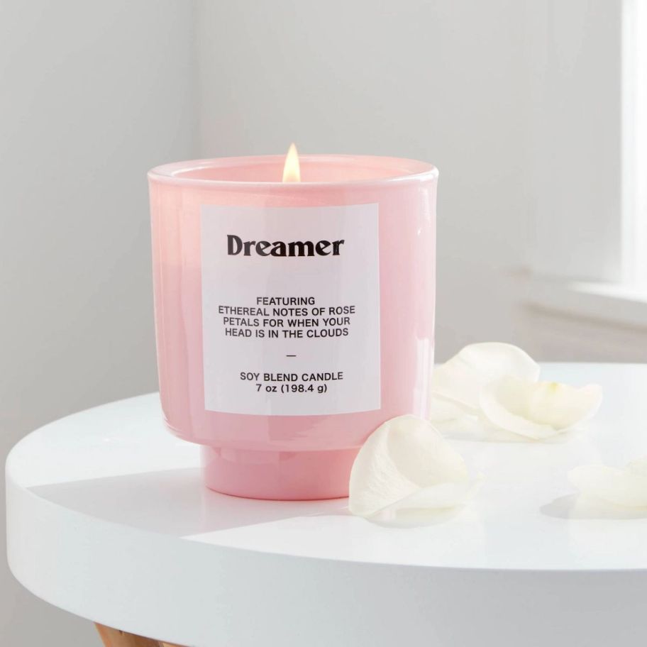 a pink jar candle lit on a tabletop next to white flower petals