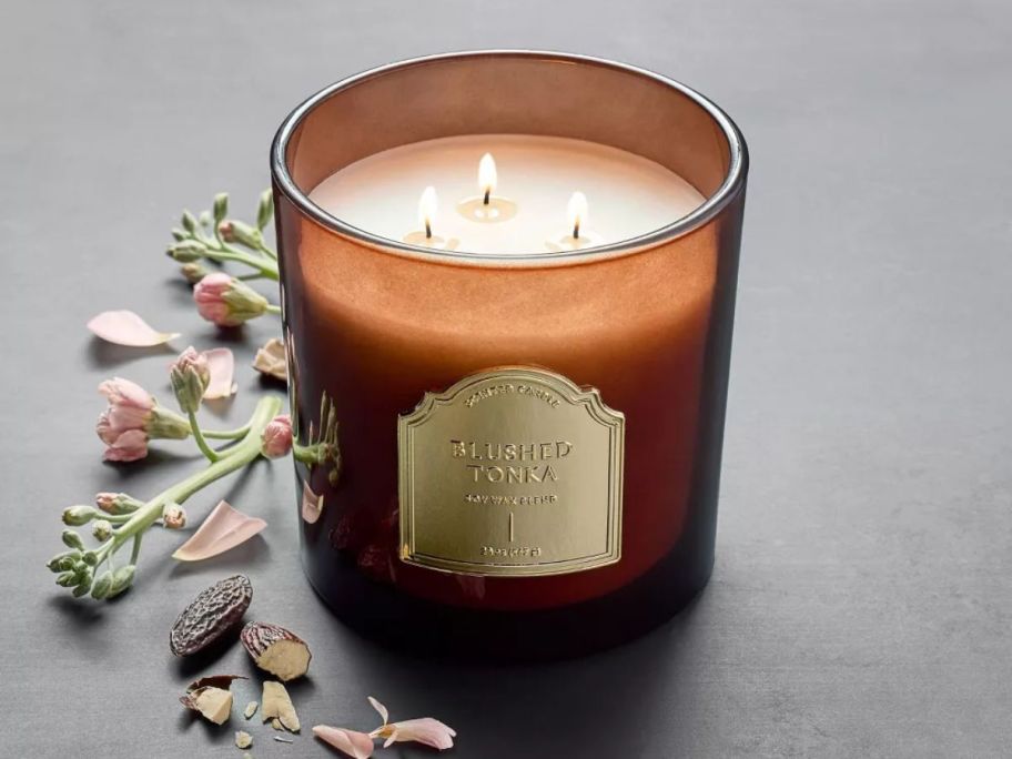 a 3 wick jar candle lit on a tabletop next to dried herbs and flowers