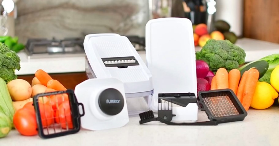 clear, white, and black vegetable chopper, slicer, grater on a kitchen counter surrounded by fruit and veggies