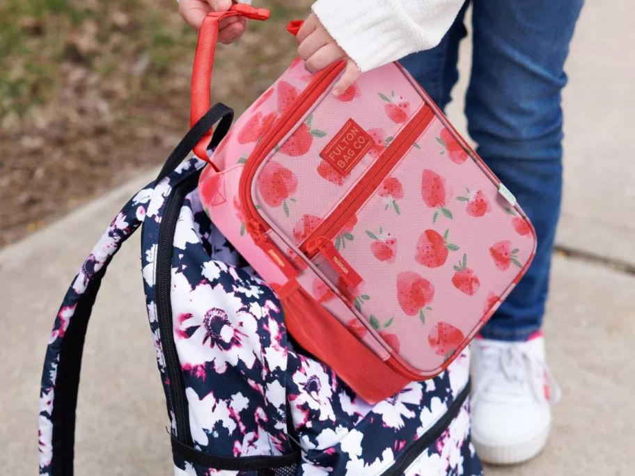 kid attaching fulton lunch bag with strawberry pattern to her backpack on sidewalk
