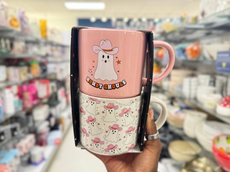 hand holding a set of 2 coffee mugs with pink ghosts wearing cowgirl hats