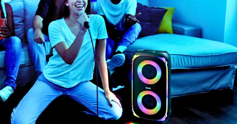 girl singing into in a microphone connected to a large bluetooth speaker with LED lights, people behind her listening