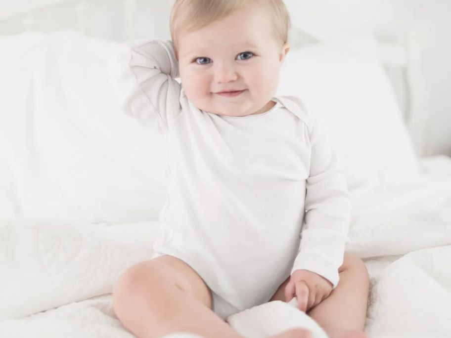 A b aby sitting on a bed wearing a Burt's Bees long sleeve white onesie