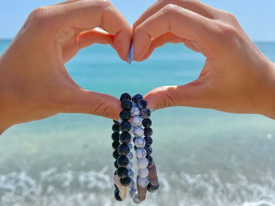 hands making a heart in front of the ocean with 3 Fahlo animal tracking bracelets dangling at the bottom of the heart