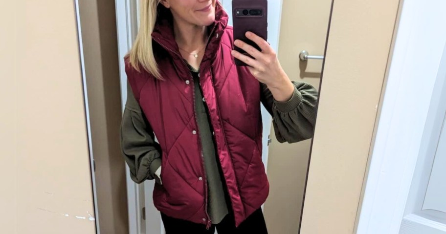 woman taking a mirror selfie wearing a wine red puffer vest with an olive green top and blue jeans