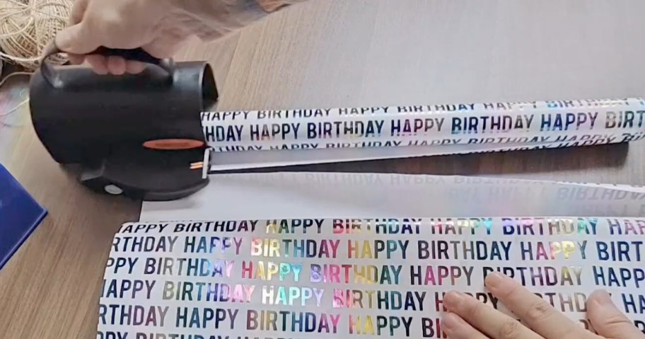 hand on a black and orange wrapping paper cutter cutting through birthday wrapping paper, more wrapping paper behind it