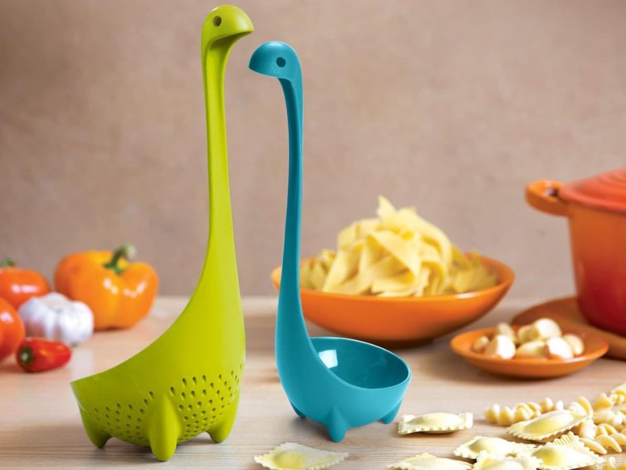 a green Nessie shaped kitchen colander ladle and smaller blue regular ladle on a kitchen counter with crackers and other food