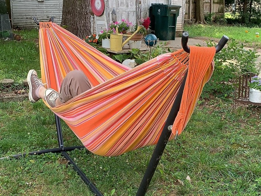 a person laying in a bright orange striped hammock on a black hammock stand in a backyard