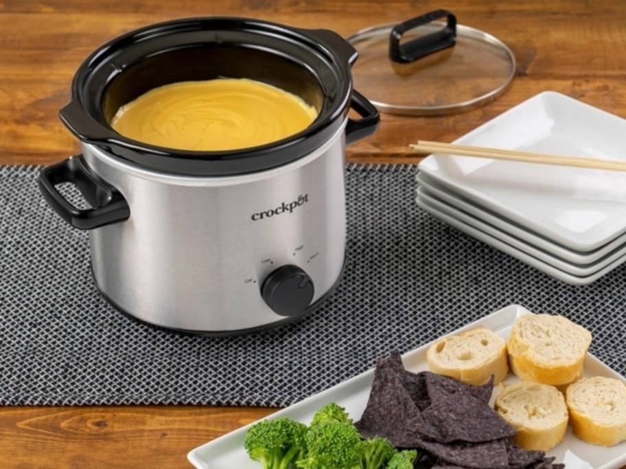 a small stainless steel and black crock pot with cheese fondue in it next to a plate of food