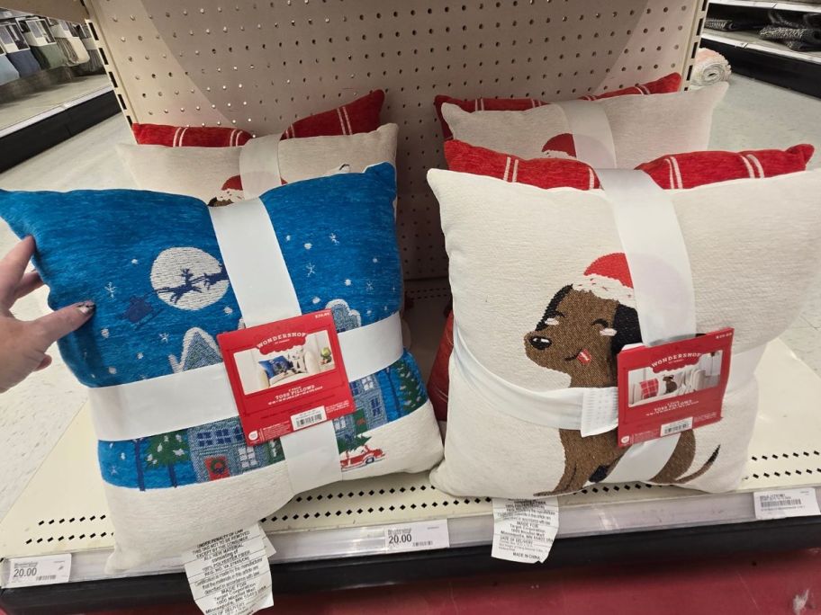 a womans hand holding a holiday throw pillow with a winter scene on it next to one with a christmas puppy on it. 