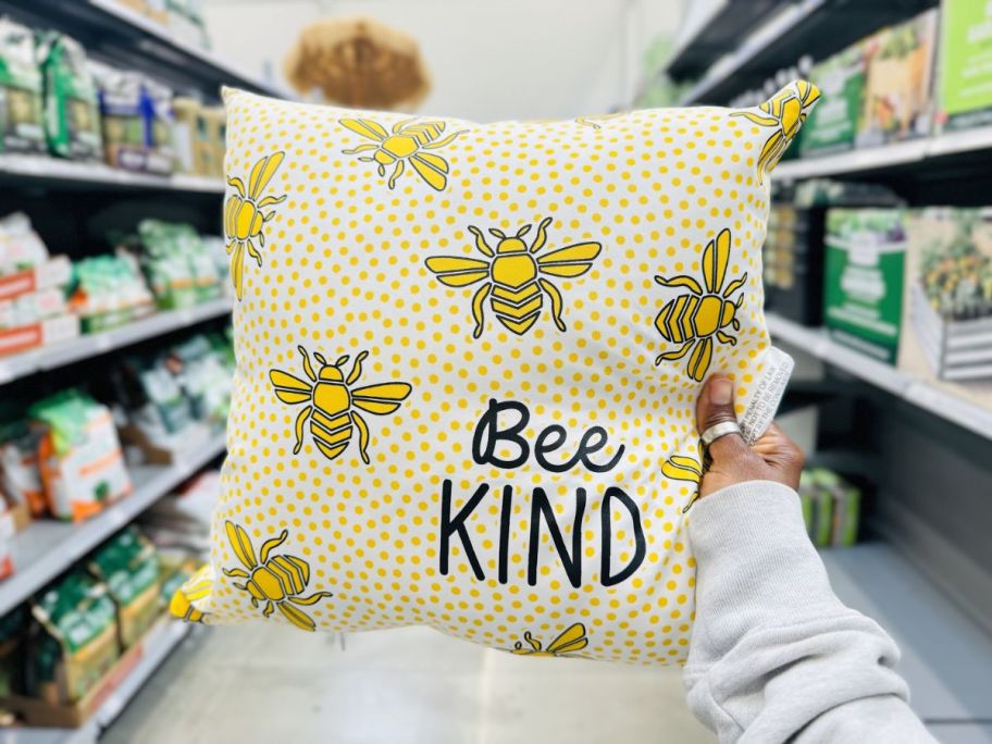 a womans hand holding up a Bee Kind throw pillow on a store aisle