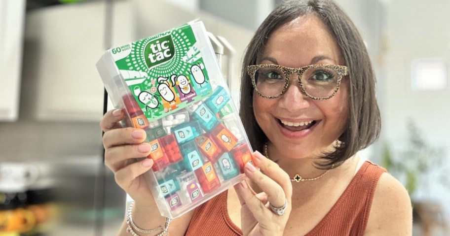 woman with brown hair and glasses smiling at the camera holding a giant Tic Tac candies box with mini Tic Tac containers inside