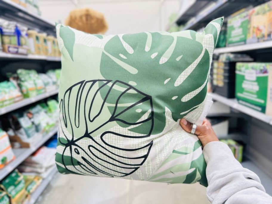 a womans hand holding up an monstera leaf throw pillow on a store aisle