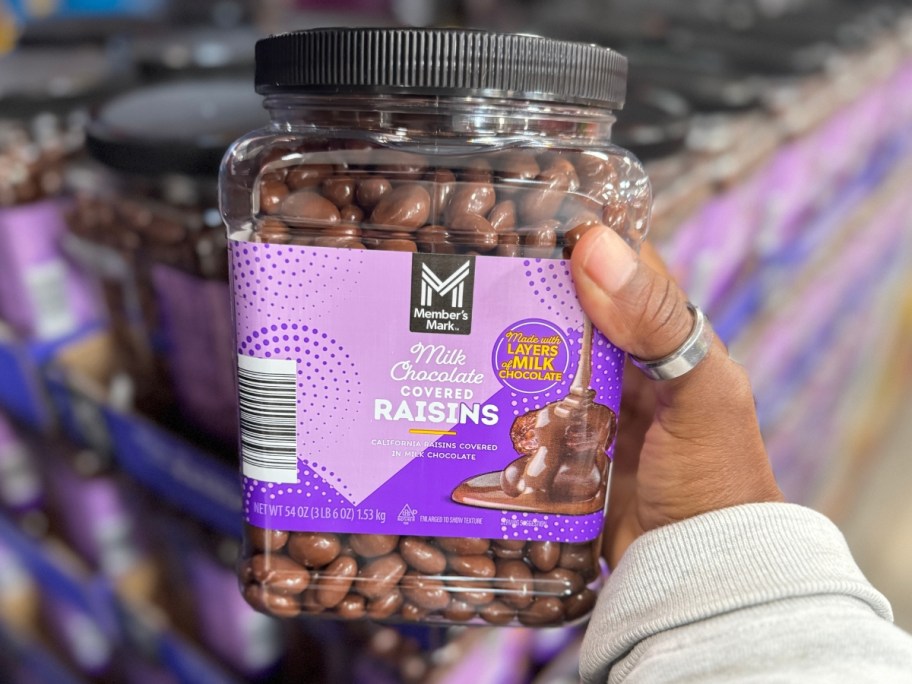 hand holding a large container with a purple label with Member's Mark Milk Chocolate Raisins in it, more on the shelf behind it